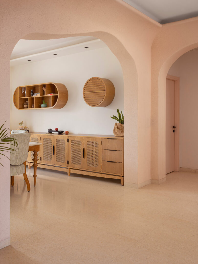 A modern dining area with light wood furniture, open shelving, decorative objects, and soft peach-colored walls with arched doorways.