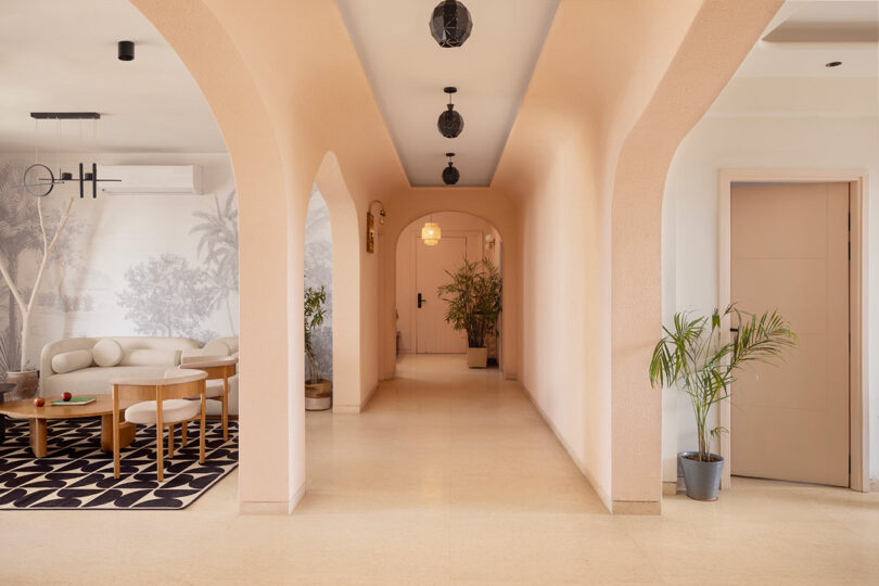 A modern hallway with peach-colored arches, potted plants, and pendant lights leads to a door; a living area with a sofa and wall mural is visible on the left.