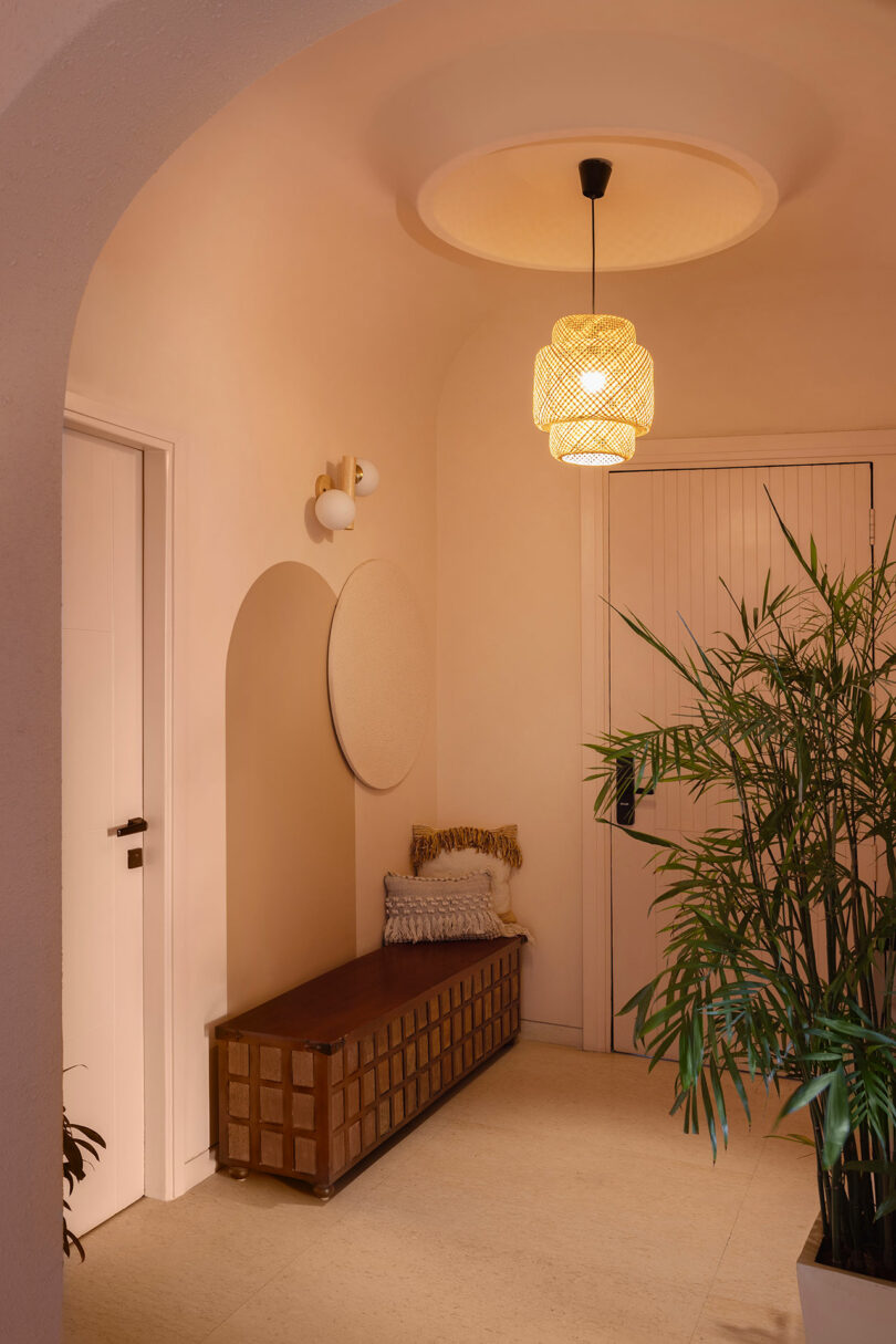 A hallway with a wooden bench, cushion, potted plant, round wall decor, wall sconce, and a pendant light hanging from a recessed ceiling.
