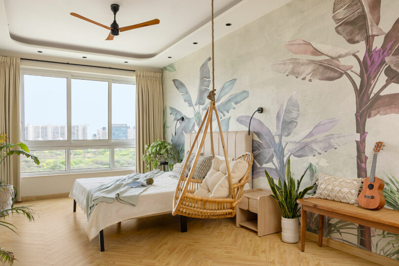 Bright bedroom with large windows, a bed, hanging chair, indoor plants, wall with tropical mural, a bench, cushions, and a small guitar.
