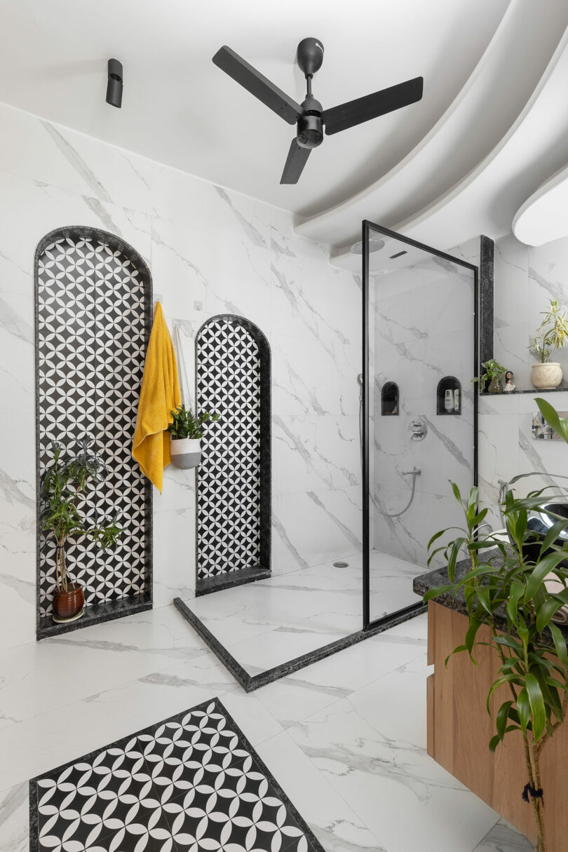 Modern bathroom with marble walls, black and white geometric tile accents, glass shower enclosure, ceiling fan, potted plants, and a yellow towel hanging on the wall.