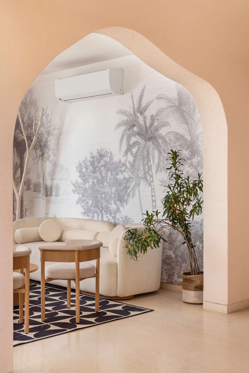 A modern living room with a beige archway, cream-colored curved sofa, wooden chairs, potted plant, and tropical wall mural, featuring an air conditioner above.