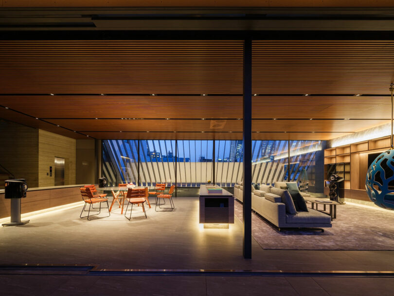 Modern, open living space with wood ceiling, glass walls, contemporary furniture, and city view at dusk. Dining area and lounge are separated by a central island.