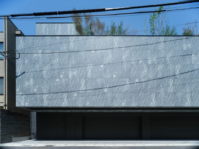 A modern building with a gray marble facade, three garage entrances, overhead utility wires, and trees visible above the roofline.