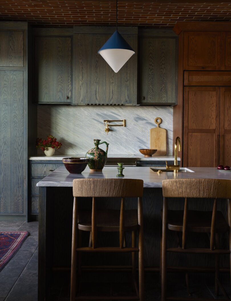 A modern kitchen with dark wood cabinets, a marble backsplash, an island with two wooden chairs, and a blue and white pendant light overhead.