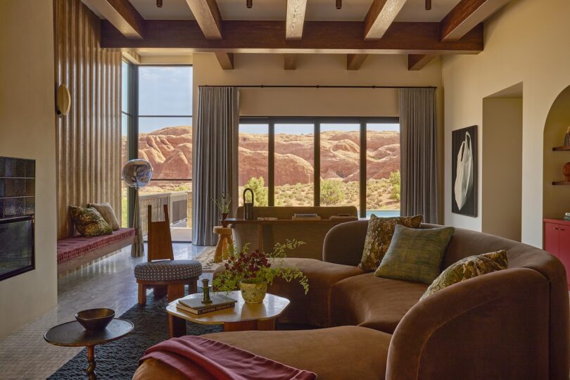 A living room with brown curved sofas, a round wooden coffee table, large windows with a desert rock view, and exposed wooden ceiling beams.