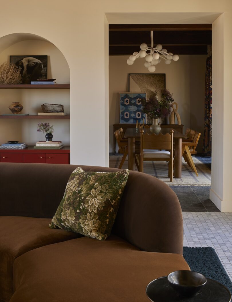 A living room with a brown curved sofa, floral pillow, built-in shelves, and a view into a dining area with a wooden table, chairs, modern chandelier, and framed artwork.