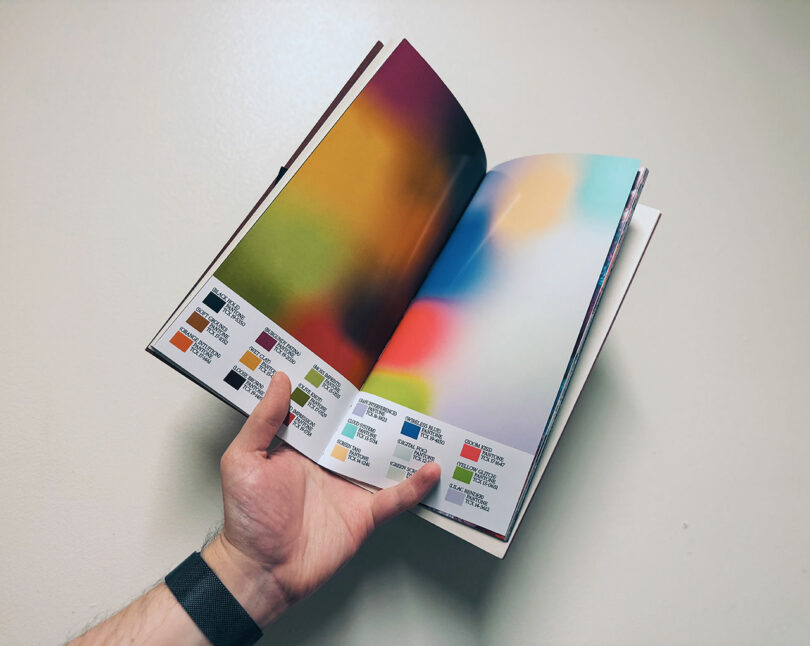 A hand holds open a book displaying abstract, blurred color images on the pages, with small color swatches and corresponding text at the bottom.