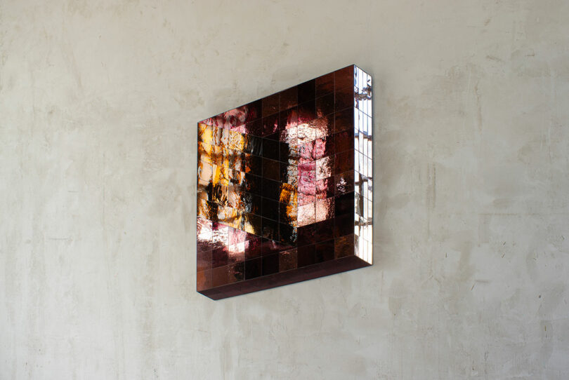 A square wall-mounted artwork made of small, reflective, reddish-brown tiles displays abstract light and color patterns against a plain, textured wall.