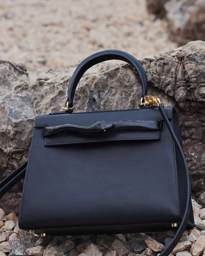 A black leather handbag with a top handle and gold hardware is placed on rocky ground in front of large stones.