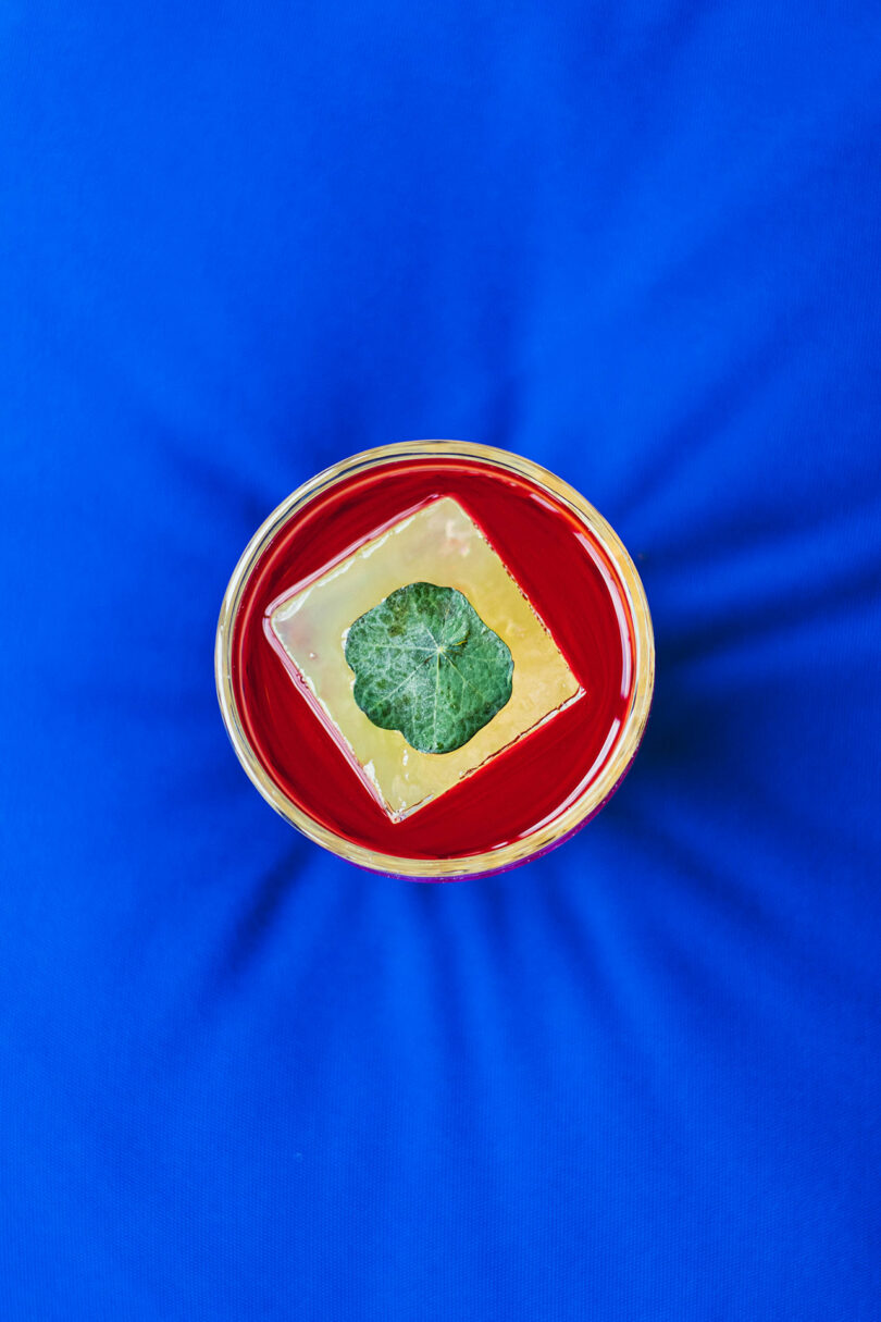A glass with a red drink, topped with a large clear ice cube and a single green leaf, viewed from above on a bright blue surface.