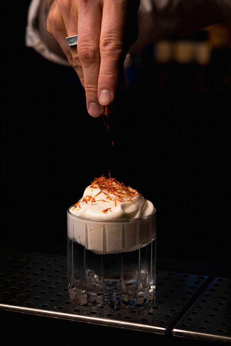 A hand sprinkles orange garnish onto a glass of dark beverage topped with a thick layer of foam.