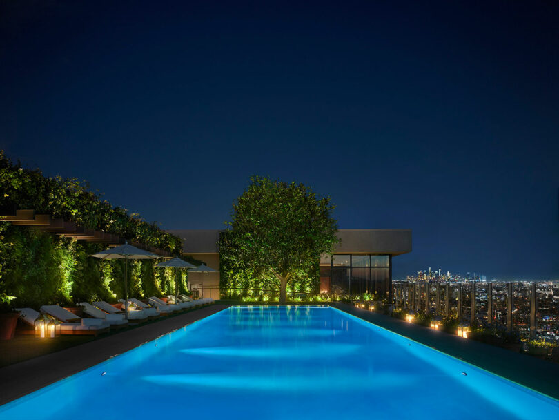 A rooftop swimming pool at night, surrounded by lounge chairs and greenery, overlooking a city skyline in the distance.