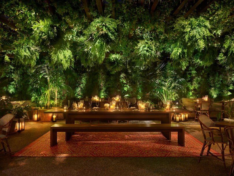 A long wooden dining table set with candles and tableware sits under lush green foliage and string lights, with rattan chairs and warm ambient lighting.