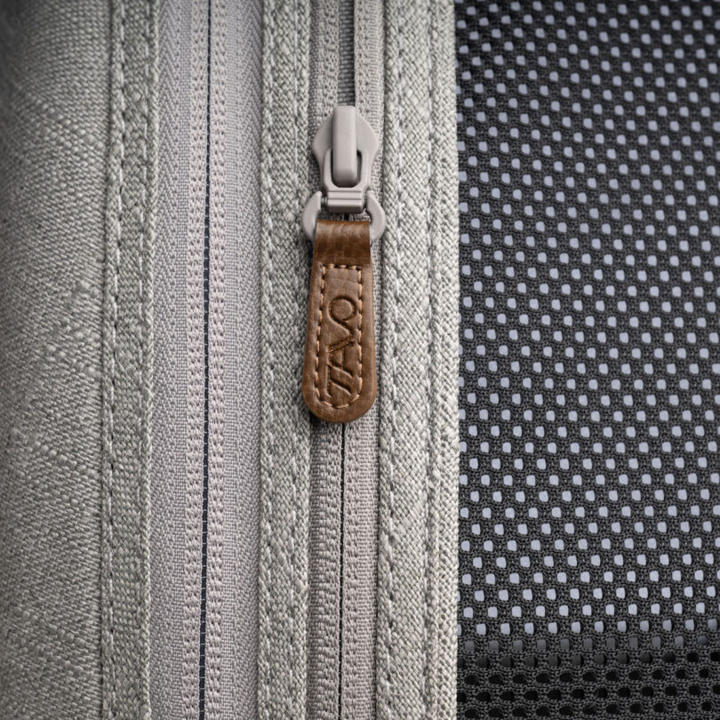 Close-up of a gray zipper with a brown, branded pull tab on a fabric background featuring a mesh section on the right