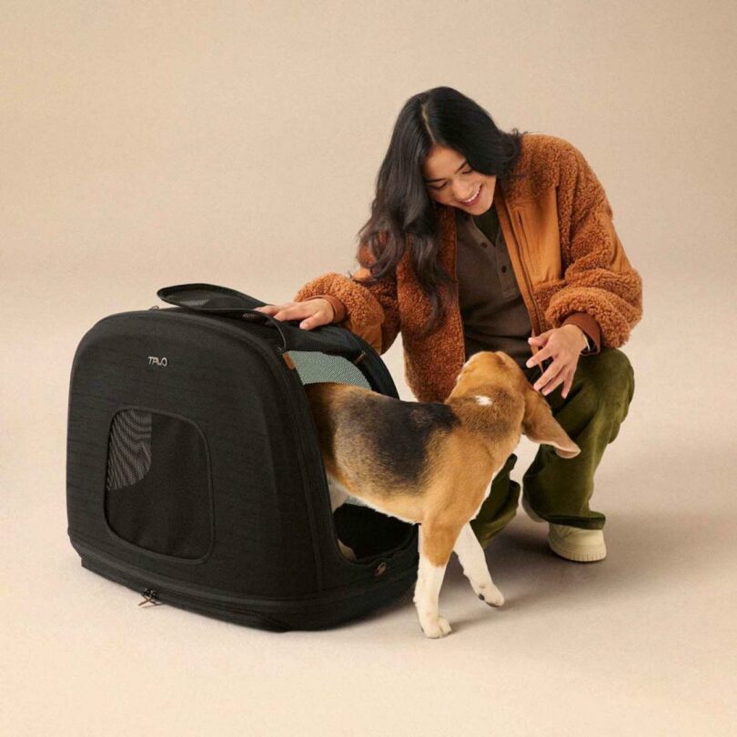 A woman in a brown jacket is smiling while helping a beagle enter a black pet carrier in a neutral-colored room