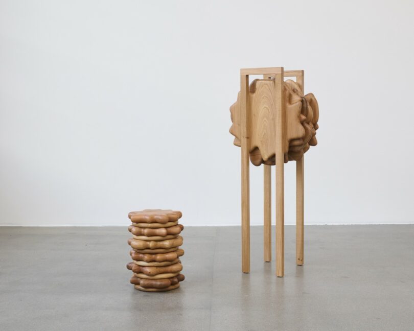 Two abstract wooden sculptures on a gray floor: a short, stacked piece on the left and a taller, four-legged structure with an organic, cloud-like center on the right