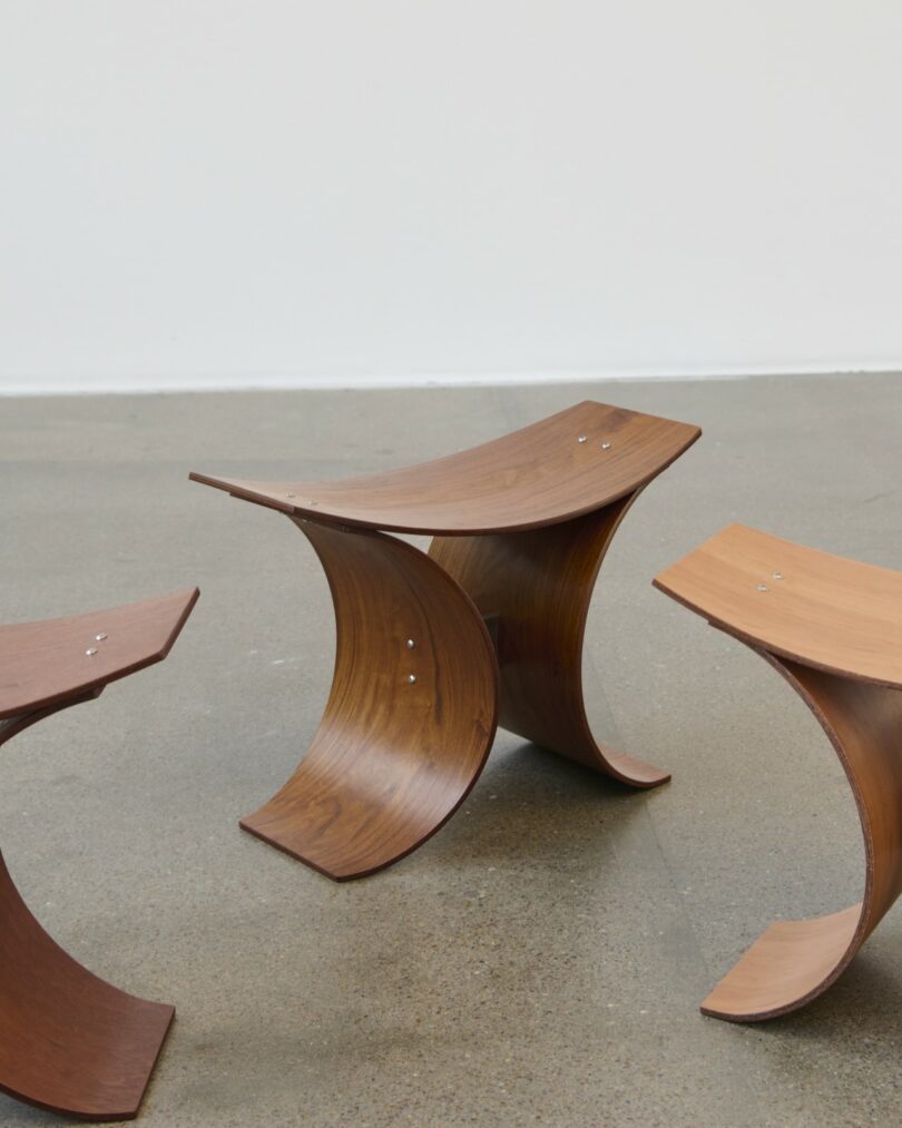 Three wooden stools with curved legs and seats are arranged on a smooth, gray floor against a plain white wall