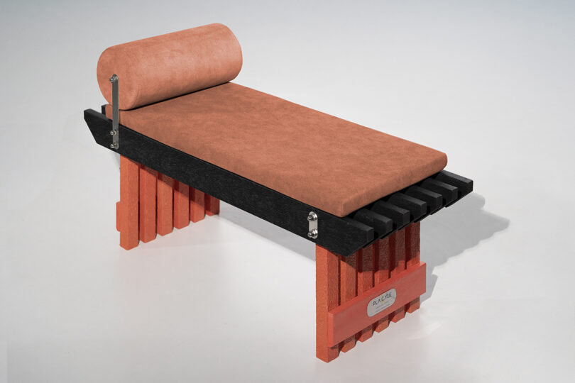 A modern bench with a rust-colored cushioned seat and cylindrical headrest, black slatted frame, and orange block-style legs.