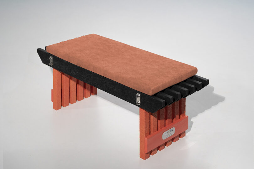A modern bench with a rectangular orange cushioned seat, black slatted frame, and orange block-style legs on a light background.