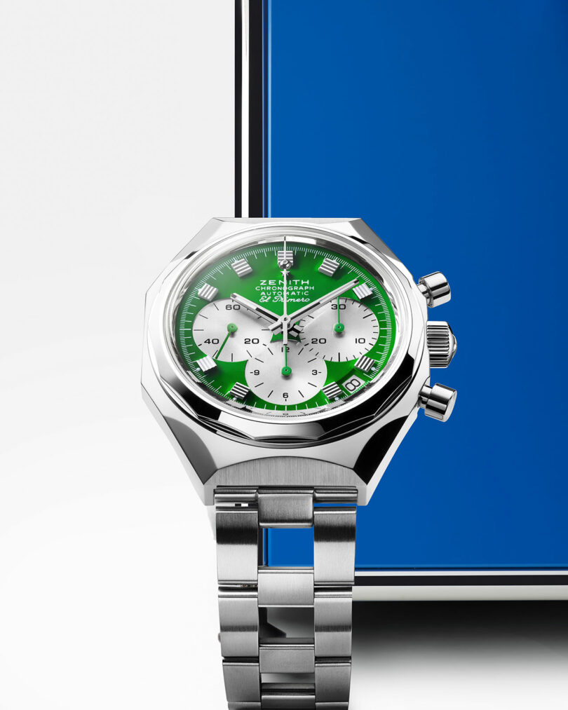 A stainless steel wristwatch with a green and white dial, three subdials, and a metal bracelet is positioned in front of a blue rectangular object.