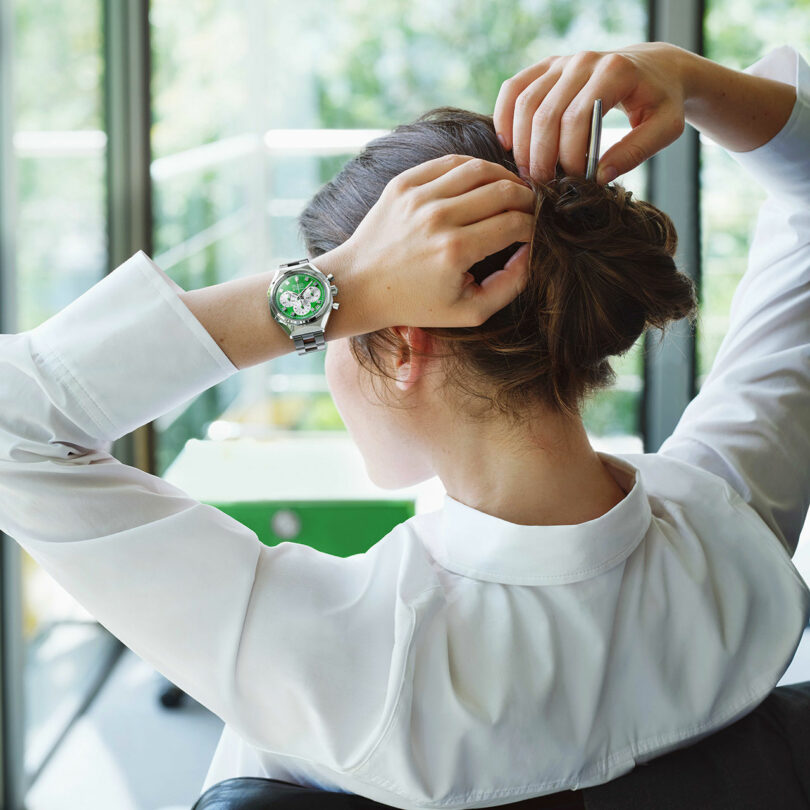 A woman in a white blouse sits in front of large windows, tying her hair up with both hands. She is wearing a silver wristwatch with a green face.