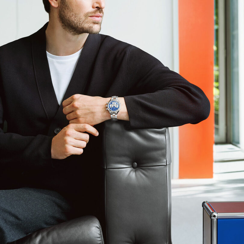 A man in a black jacket and white shirt sits on a leather chair, resting his arm on the chair back and wearing a silver wristwatch with a blue face.
