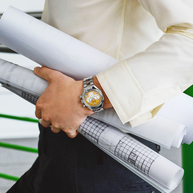 A person wearing a light shirt and wristwatch holds several rolled-up architectural blueprints under their arm.