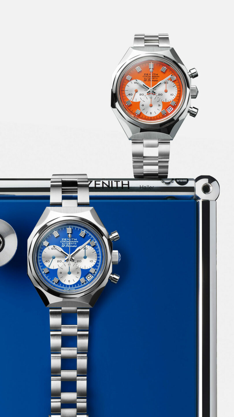 Two stainless steel chronograph watches with colored dials—one orange, one blue—are displayed on a metallic frame against a white background.
