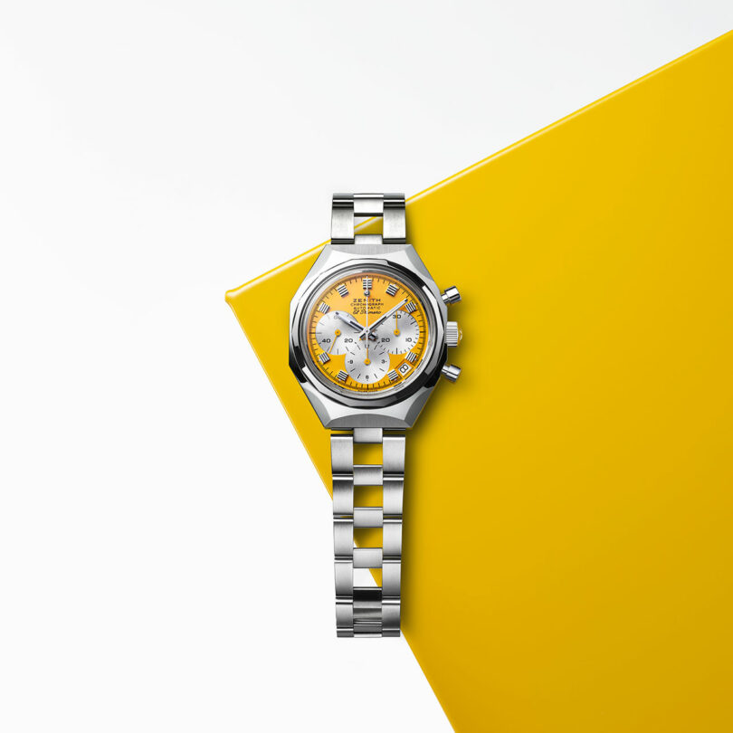 A stainless steel wristwatch with a yellow and white dial is displayed on a yellow geometric surface against a white background.