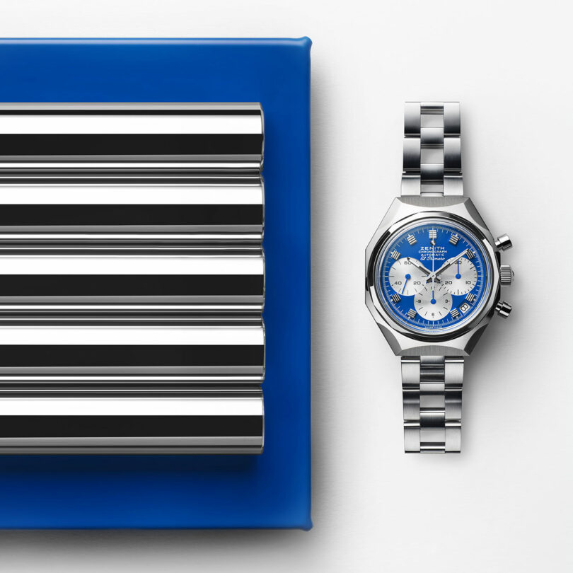 A silver chronograph wristwatch with a blue and white dial sits next to a blue tray holding four parallel steel cylinders on a white surface.