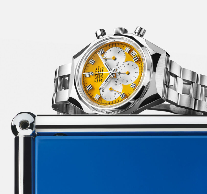 A silver wristwatch with a yellow and white dial rests on top of a shiny blue rectangular object against a white background.