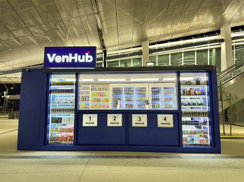 A VenHub automated store kiosk at night, displaying various snacks, drinks, and other convenience items behind glass, with four numbered pick-up windows.