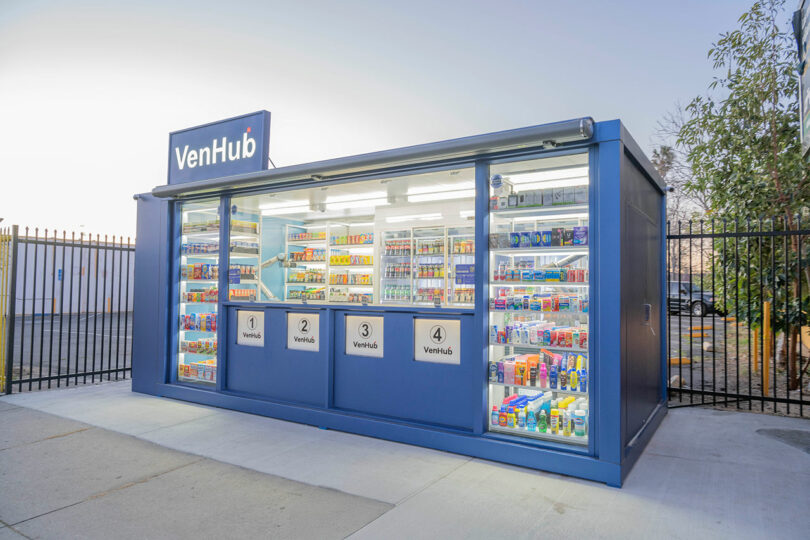 A blue VenHub vending kiosk stocked with snacks, drinks, and everyday items stands outdoors near a fence and trees.