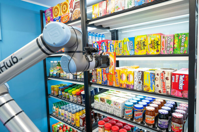 A robotic arm selects an item from a grocery shelf stocked with various cereals, snacks, sauces, and beverages in a brightly lit store.