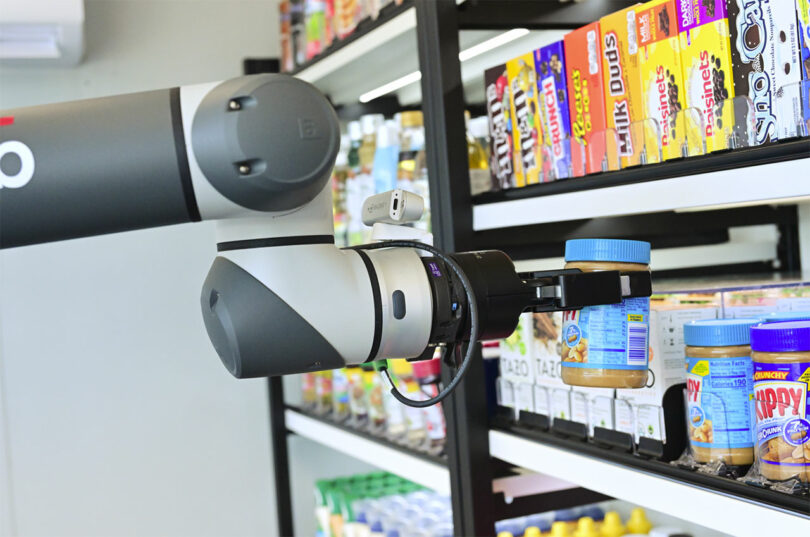 A robotic arm selects a jar of peanut butter from a supermarket shelf lined with various food items.