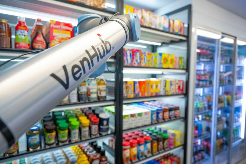 A close-up of a VenHub device in a brightly lit store aisle with shelves stocked with packaged food, drinks, and refrigerated items.