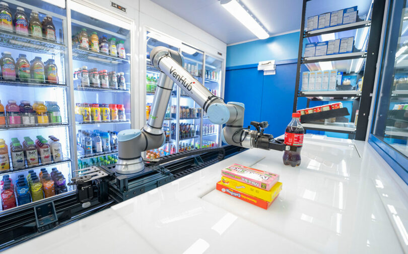 A robotic arm picks up a bottle of soda near grocery shelves stocked with drinks and boxes of food on a white counter.