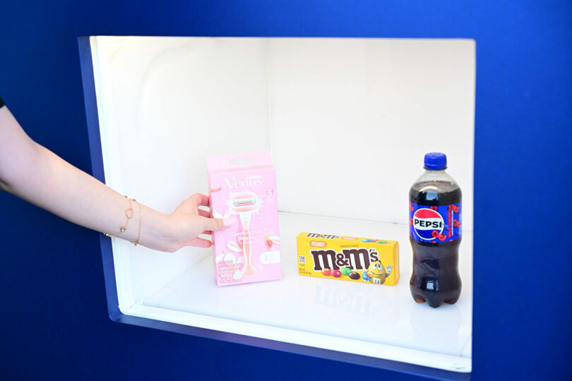 A hand reaching for a pink Venus razor pack next to a yellow box of M&M's and a bottle of Pepsi inside a brightly lit display case.