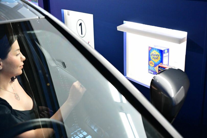 A woman in a car at a drive-thru window receives a box of Honey Maid graham crackers from a shelf.