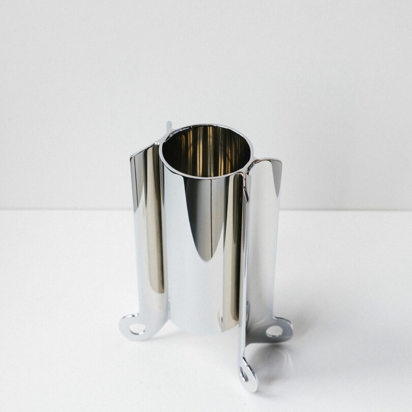 A polished metal cylindrical object with three curved extensions and circular cutouts, placed on a white surface against a plain background.