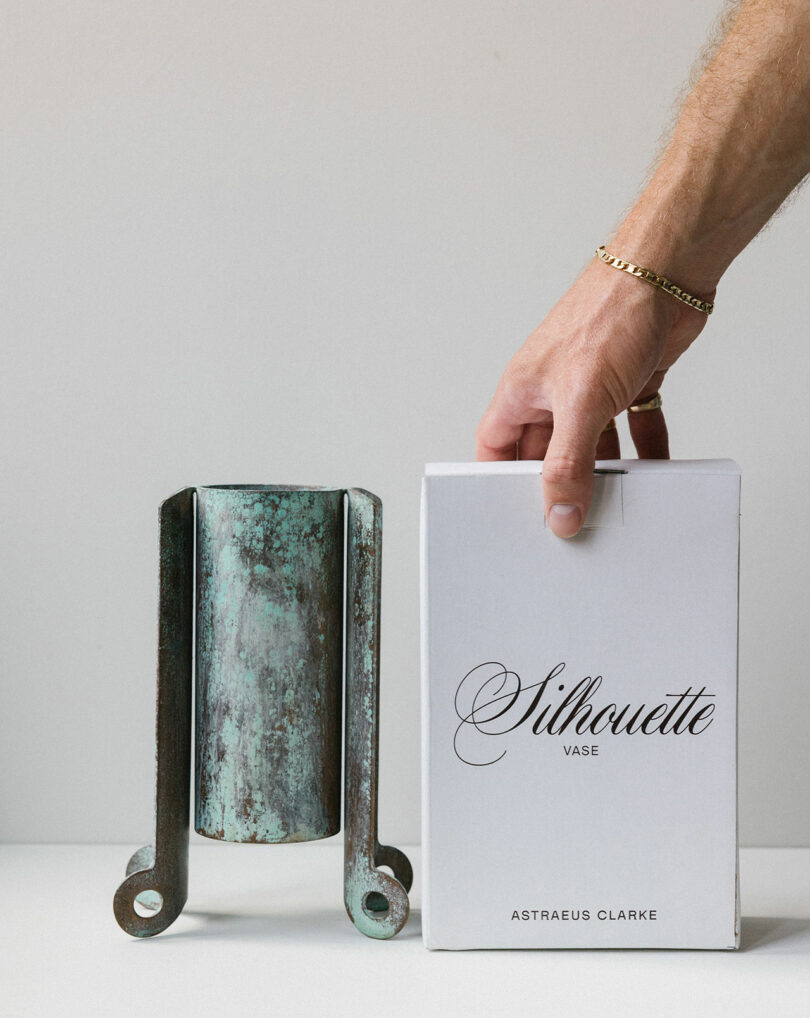 A hand places a white box labeled "Silhouette Vase Astraeus Clarke" beside a metal vase with a rustic, oxidized finish on a white surface.