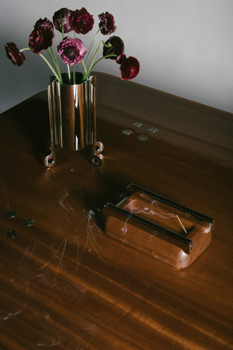 A metallic vase with dark red flowers, a wooden box with smoke, and several coins are arranged on a polished brown table.