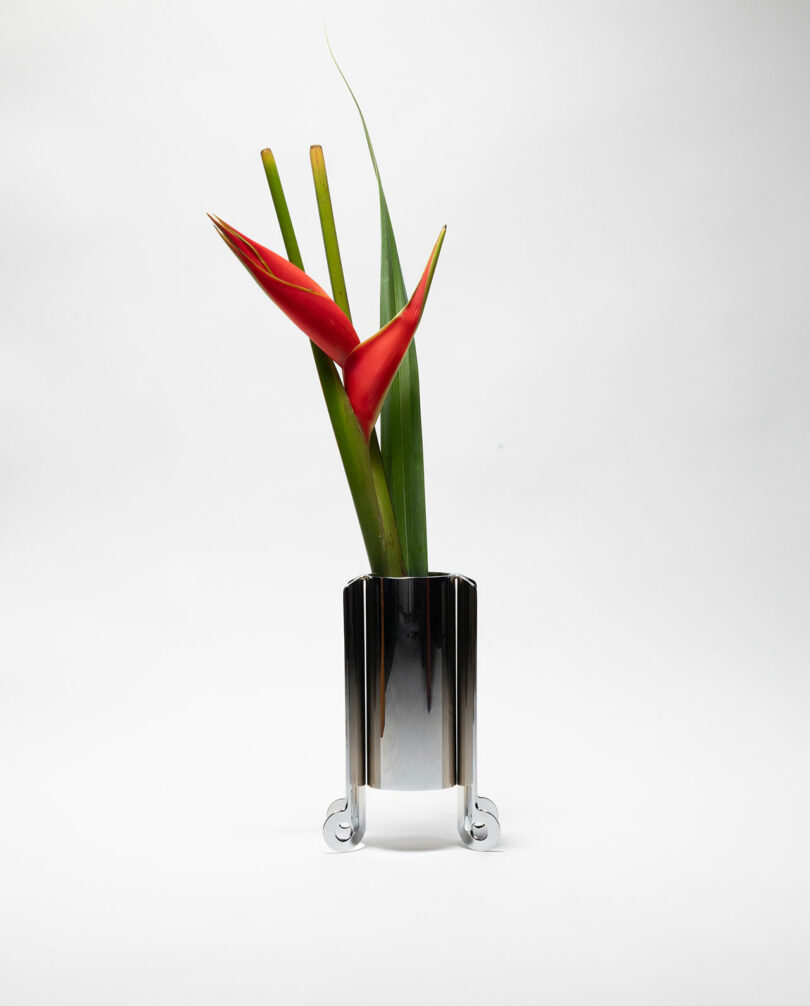 A shiny metal vase with three green leaves and two red tropical flowers, set against a plain white background.