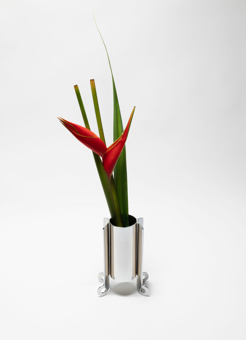 A metallic vase with a modern design holds two red heliconia flowers and green leaves against a plain white background.