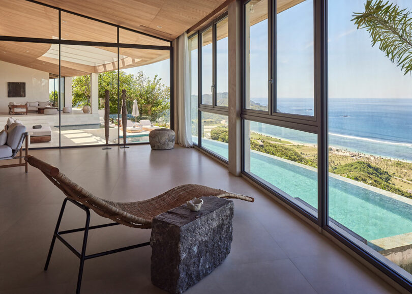 Modern living space with large windows overlooking an infinity pool and ocean view, featuring minimalist furniture and natural materials.