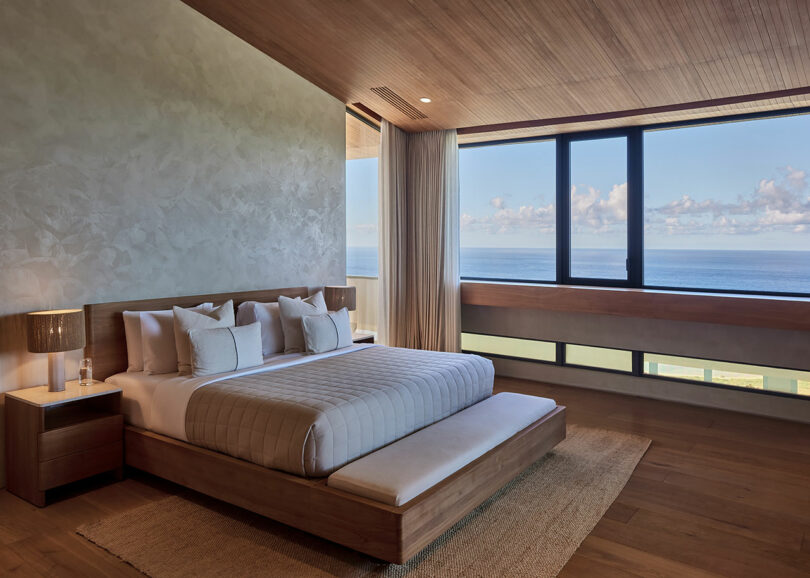 Modern bedroom with a wooden bed, neutral decor, large window, ocean view, two nightstands with lamps, and natural light.