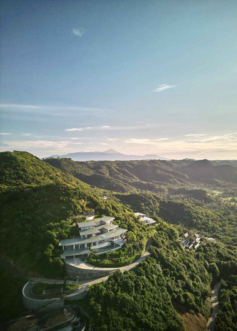 A modern house with curved architecture is nestled among green hills under a clear sky, with distant mountains visible on the horizon.