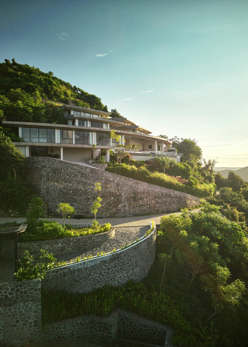 Modern hillside villas with large windows and stone retaining walls overlook a lush, green landscape under a clear sky.