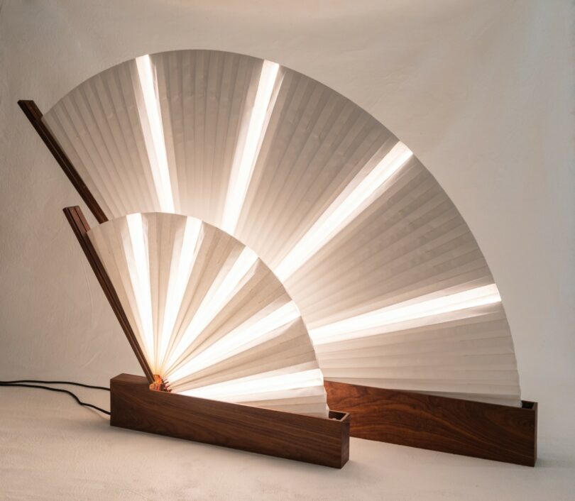 Two decorative lamps shaped like folding fans with wooden bases and illuminated pleated shades, positioned against a plain background